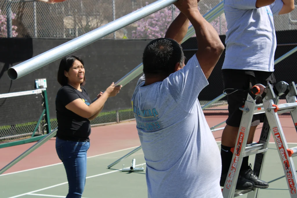 Team of one female and two males setting up large tent on tennis court, adhering to safety protocols.