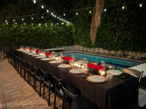 Elegant nighttime poolside dinner setup with string lights, black chairs, formal place settings, and red floral centerpieces—ideal for intimate party event rentals in Los Angeles.
